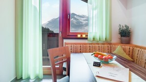 Uma área de refeições com uma mesa, cadeiras, uma tigela de frutas, um cardápio e vista para montanhas pela janela.