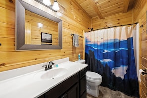 A wooden cabin bathroom with a white sink, a mirror, and a shower curtain depicting a mountain landscape. - Theater · Game Room · Hot Tub · Dog-Friendly · Sleeps 12 (Sevierville)