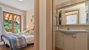 Quarto com cama, banheiro com pia e espelho, e vista para o verde lá fora.