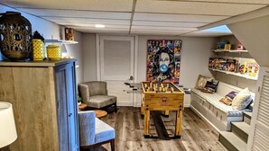 Une pièce chaleureuse avec un baby-foot, un canapé, un fauteuil et des étagères remplies de jeux de société.