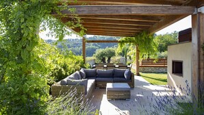 An outdoor patio with a wooden pergola, a grey sofa, and a stone bench. - Villa San Francesco - Homelike Villas (Assisi)