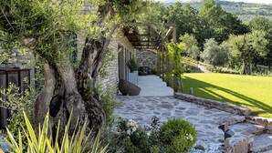 A stone pathway leading to a wooden pergola with a stone wall backdrop, surrounded by greenery and a clear view of the countryside. - Villa San Francesco - Homelike Villas (Assisi)