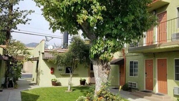 Um edifício de dois andares com fachada verde, uma escadaria à direita e uma árvore em primeiro plano.