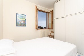A bedroom with a bed, a radiator, a wooden chair, a window with an open shutter, and a framed picture on the wall.