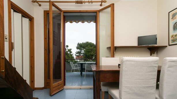 A room with a wooden staircase, a dining table with chairs, a TV on a shelf, and a view through a glass door. - M134 - Marcelli, four-room apartment with terrace 50 meters from the sea (Marcelli)