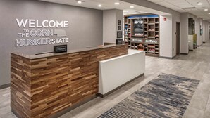 A reception desk with a 'Welcome to the Corn Husker State' sign, a Hilton Honors logo, and a bookcase with various books and magazines. - Hampton Inn Columbus NE (Columbus)