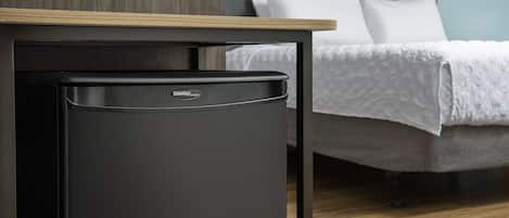 A compact black refrigerator with a Danby logo, a wooden table with a light-colored top, and a bed with white bedding in the background.