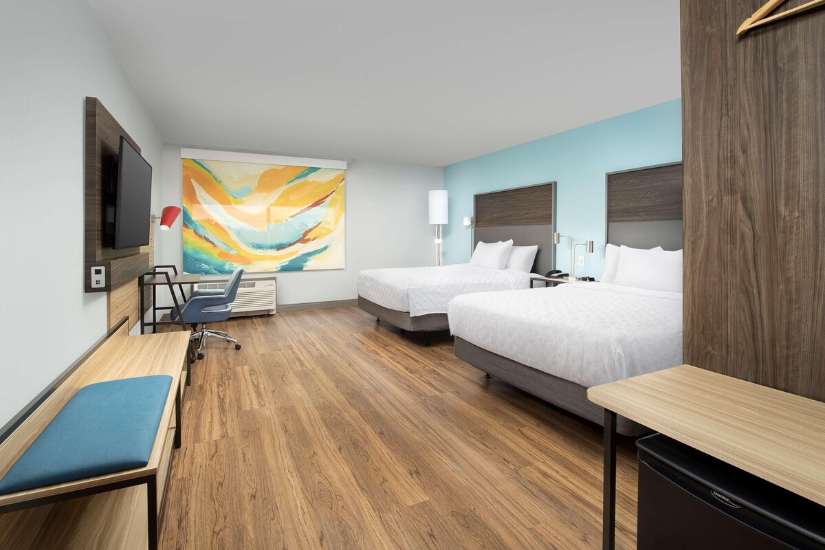 Une chambre d’hôtel avec deux lits, un bureau, une chaise, une télévision et un grand tableau abstrait accroché au mur.