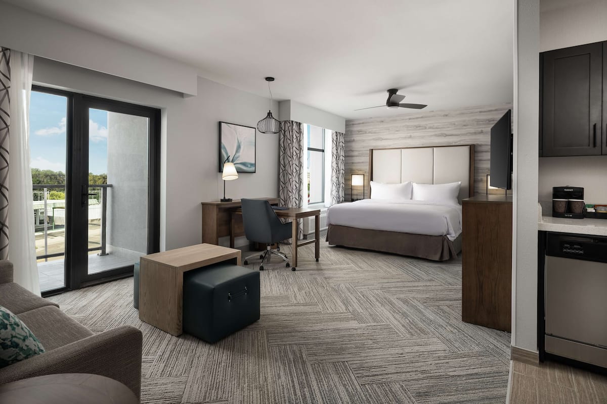 A modern hotel room with a large bed, a desk, a chair, a sofa, a dining table, and a kitchen area.