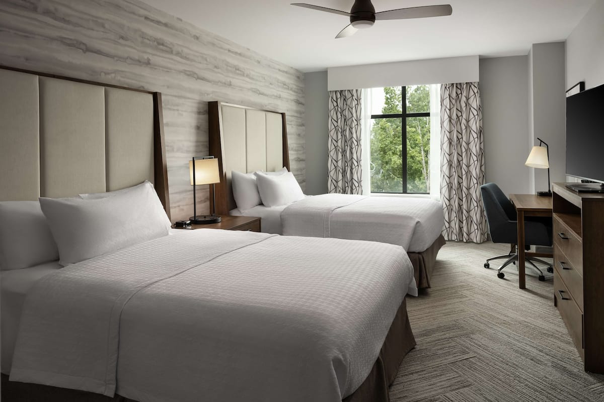 A hotel room with two beds, a desk, a chair, a TV, and a window with curtains.