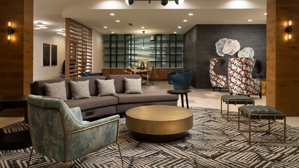 Una sala de estar moderna con un sofá modular, una mesa de centro redonda y una alfombra estampada. Hay dos sillones, una estantería y un aplique de pared.