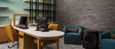 A modern office space with a desk, computer, and ergonomic chair. There are two teal armchairs and a black speaker. The room features a brick wall and a green shelving unit with decorative items.