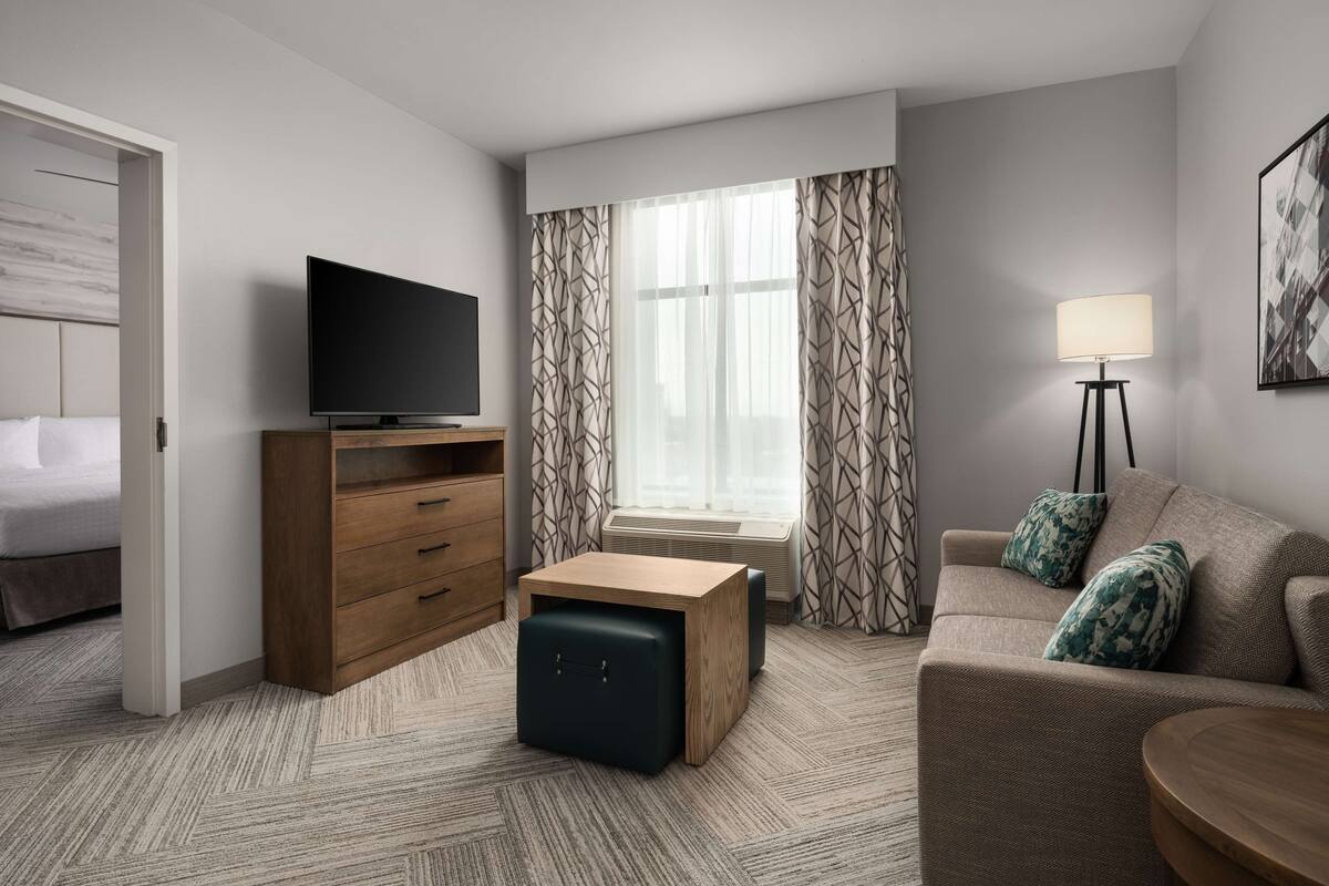 A hotel room with a bed, a TV, a wooden dresser, a sofa, a lamp, and a window with curtains.