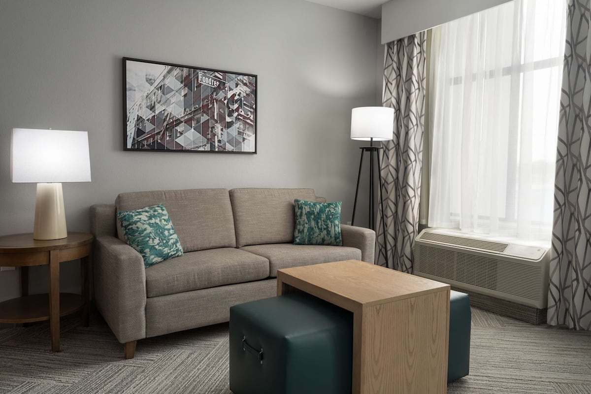 A modern hotel room with a sofa, a small table, a standing lamp, a large window with patterned curtains, and a cityscape artwork on the wall.