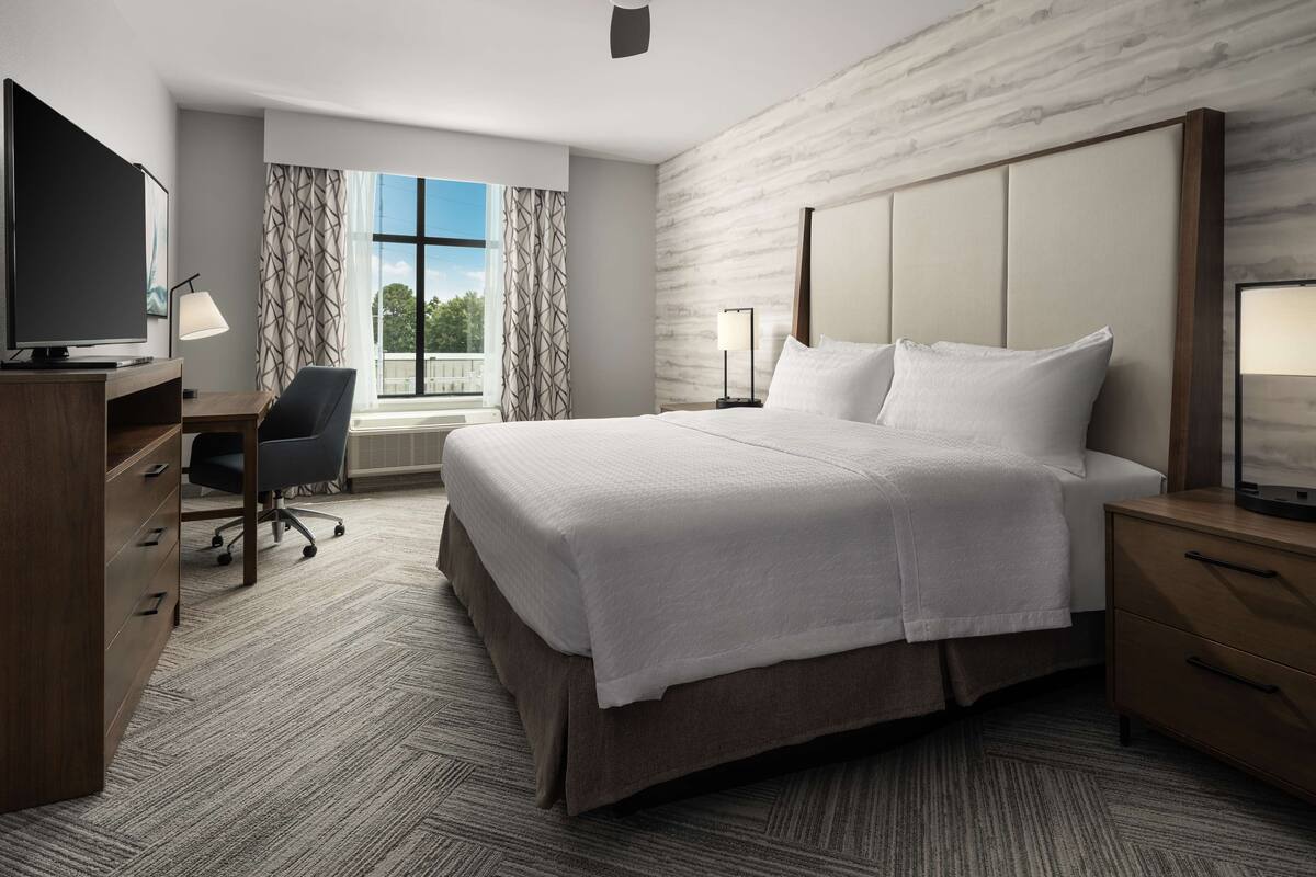 A hotel room with a bed, desk, chair, TV, and a window with curtains.