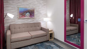 A hotel room with a sofa, a side table, a lamp, a mirror, and a patterned carpet.