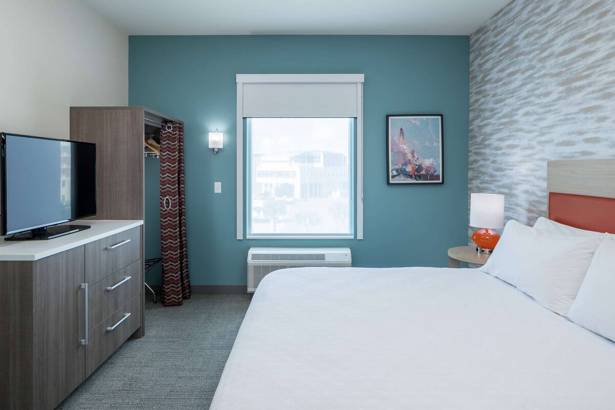 Une chambre d’hôtel avec un lit, un téléviseur posé sur une commode, une fenêtre avec des rideaux, une lampe fixée au mur et un tableau encadré.