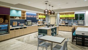 A modern cafe with a clean layout, a self-service counter, and a seating area with tables and chairs. - Home2 Suites by Hilton Cape Canaveral Cruise Port, FL (Cape Canaveral)