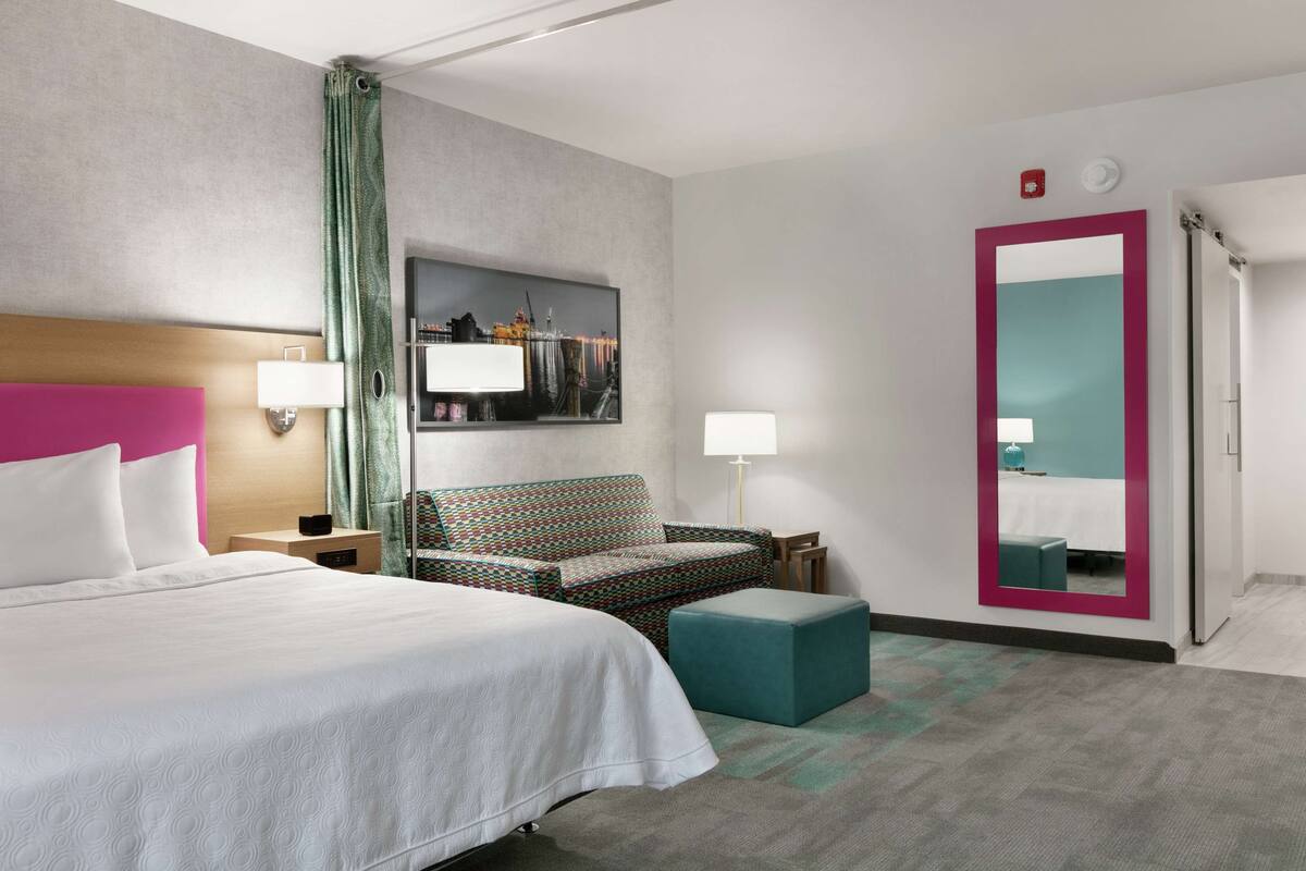 Ein Hotelzimmer mit einem großen Bett, einem gemusterten Sessel, einem kleinen Hocker, einem Spiegel und einem Stadtbild an der Wand.
