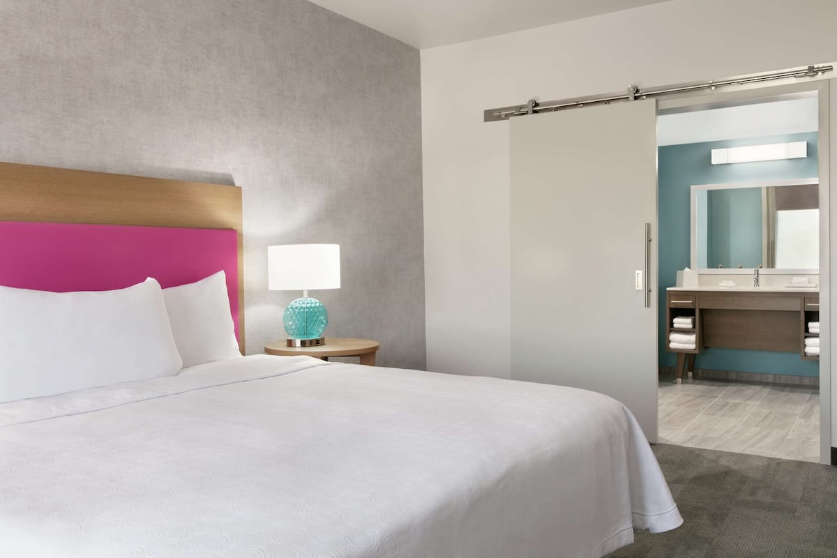 Ein Hotelzimmer mit einem großen Bett, einem Nachttisch mit Lampe und einem durch eine offene Tür einsehbarer Badezimmer.