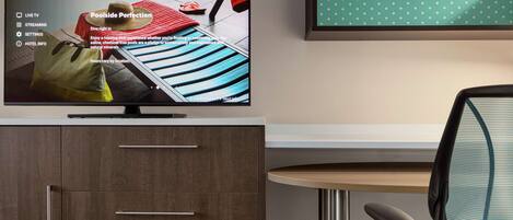 A hotel room with a TV displaying a poolside advertisement, a wooden cabinet with drawers, a green chair, and a polka dot wall decoration.