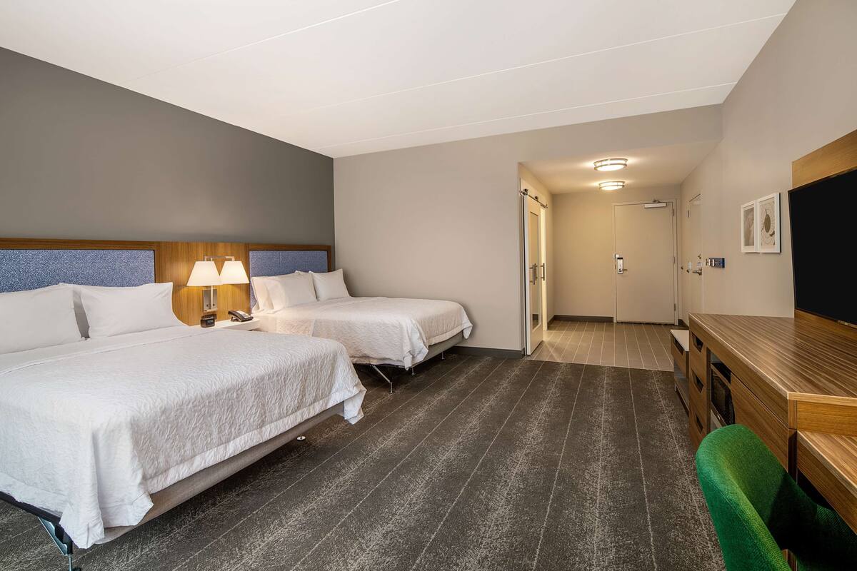A hotel room with two beds, a desk, and a chair. There is a door leading to a bathroom and a hallway.