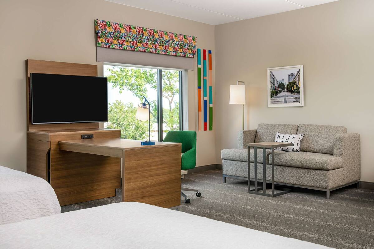 A hotel room with a wooden desk, a flat-screen TV, a green chair, a grey sofa, and a framed picture on the wall.
