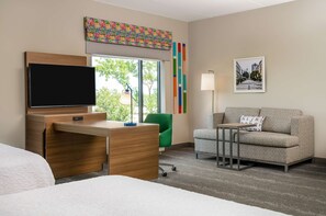 A hotel room with a wooden desk, a flat-screen TV, a green chair, a grey sofa, and a framed picture on the wall.