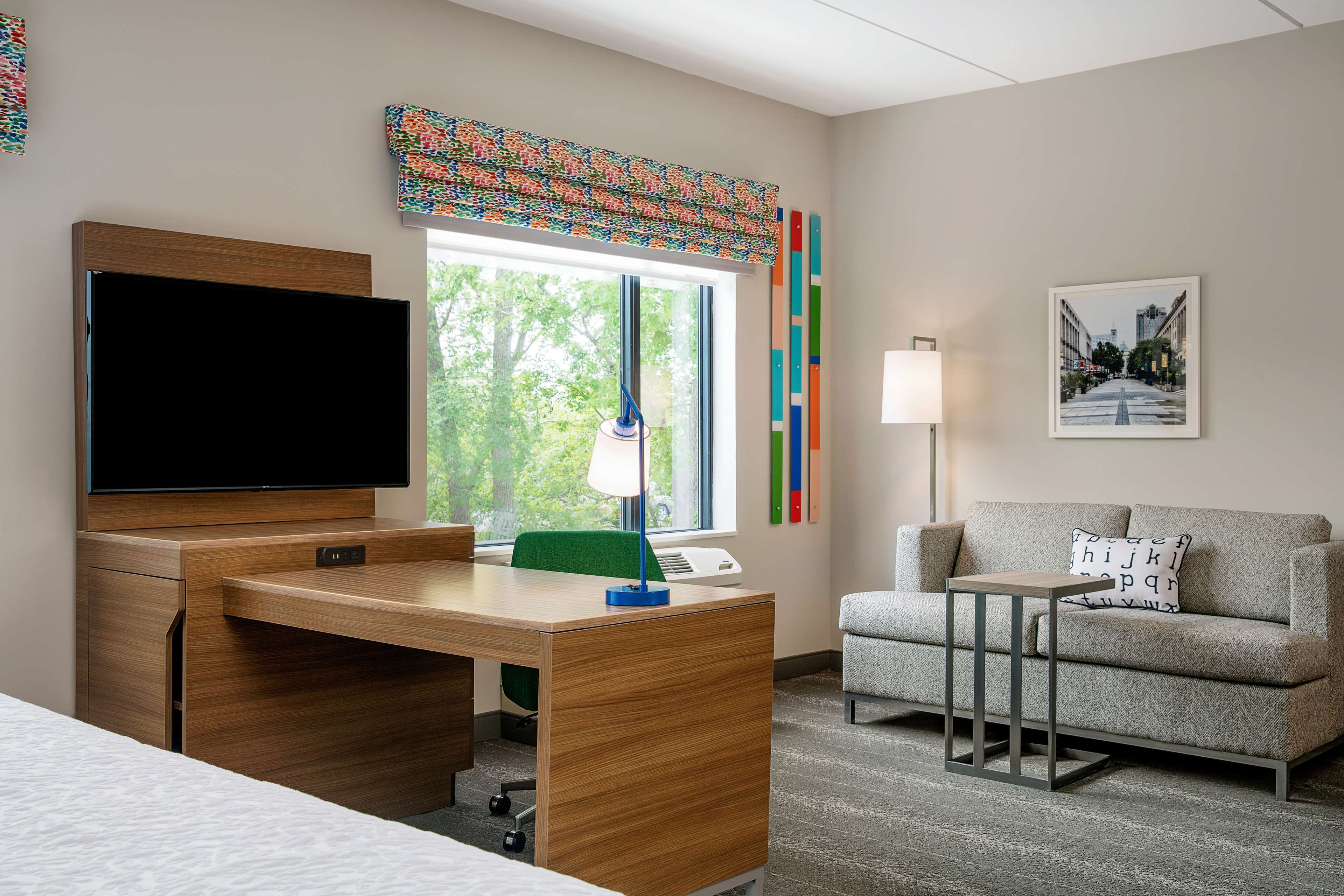 A hotel room with a wooden desk, a flat-screen TV, a green chair, a grey sofa, a small side table, and a window with a view of trees.