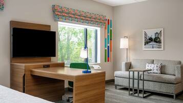 A hotel room with a wooden desk, a flat-screen TV, a green chair, a grey sofa, a small side table, and a window with a view of trees.
