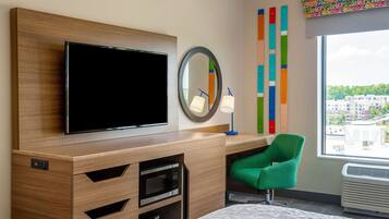 Ein Hotelzimmer mit Flachbildfernseher, Spiegel, grünem Sessel, Bett und Fenster mit Blick auf die Stadt.