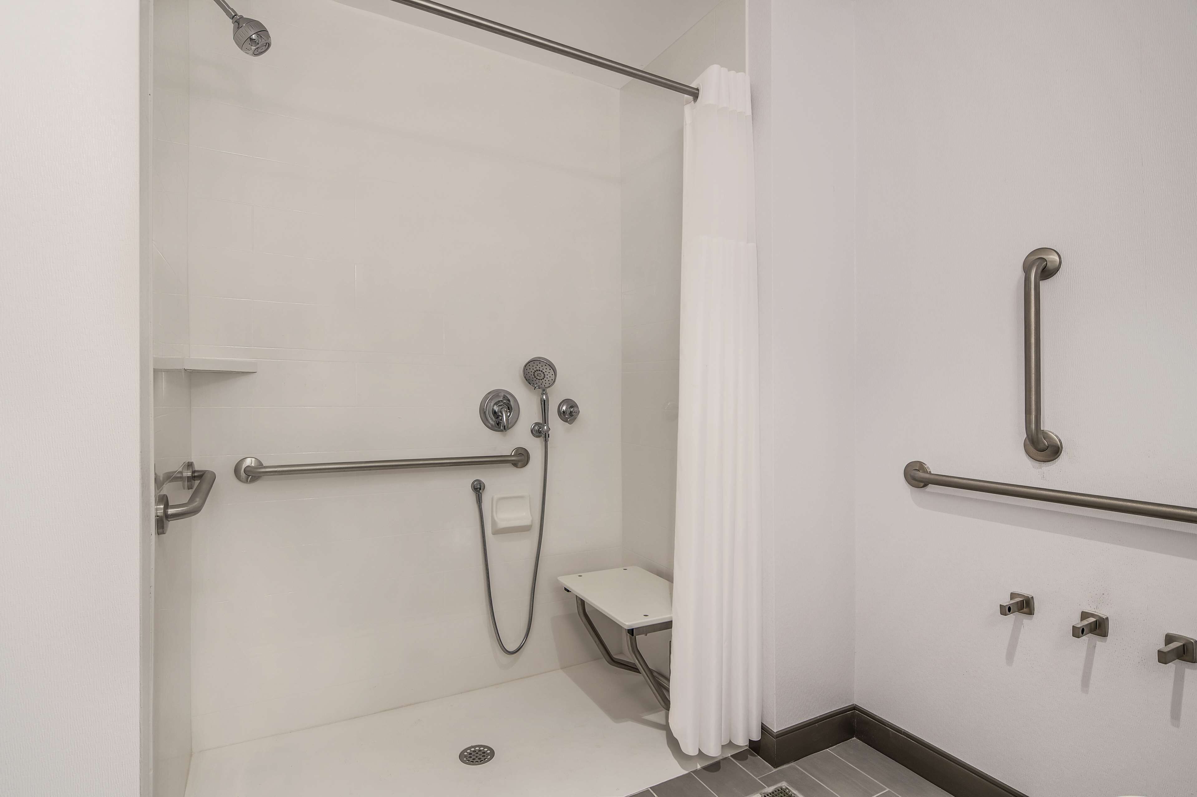 Une salle de bain avec une douche à l’italienne, des barres d’appui, des toilettes blanches et un rideau de douche.