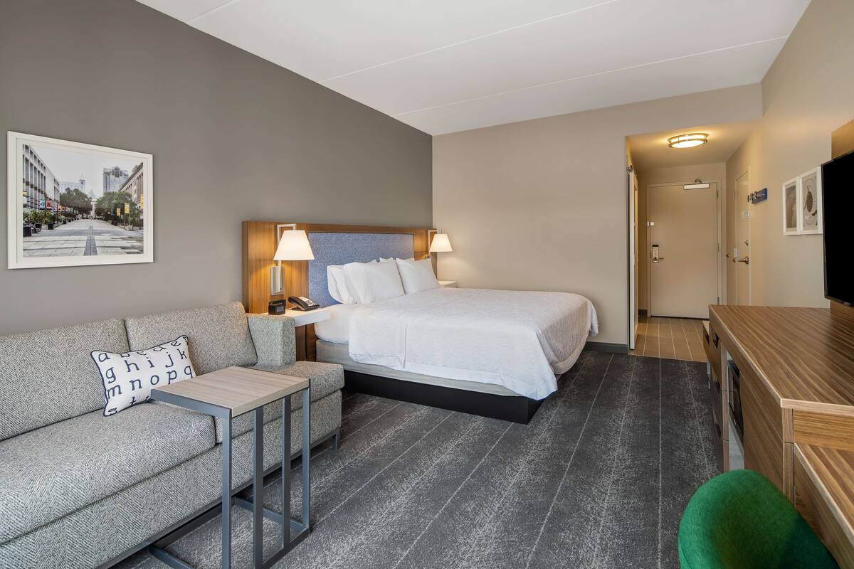 A hotel room with a bed, a sofa, a small table, a TV, and a painting on the wall.