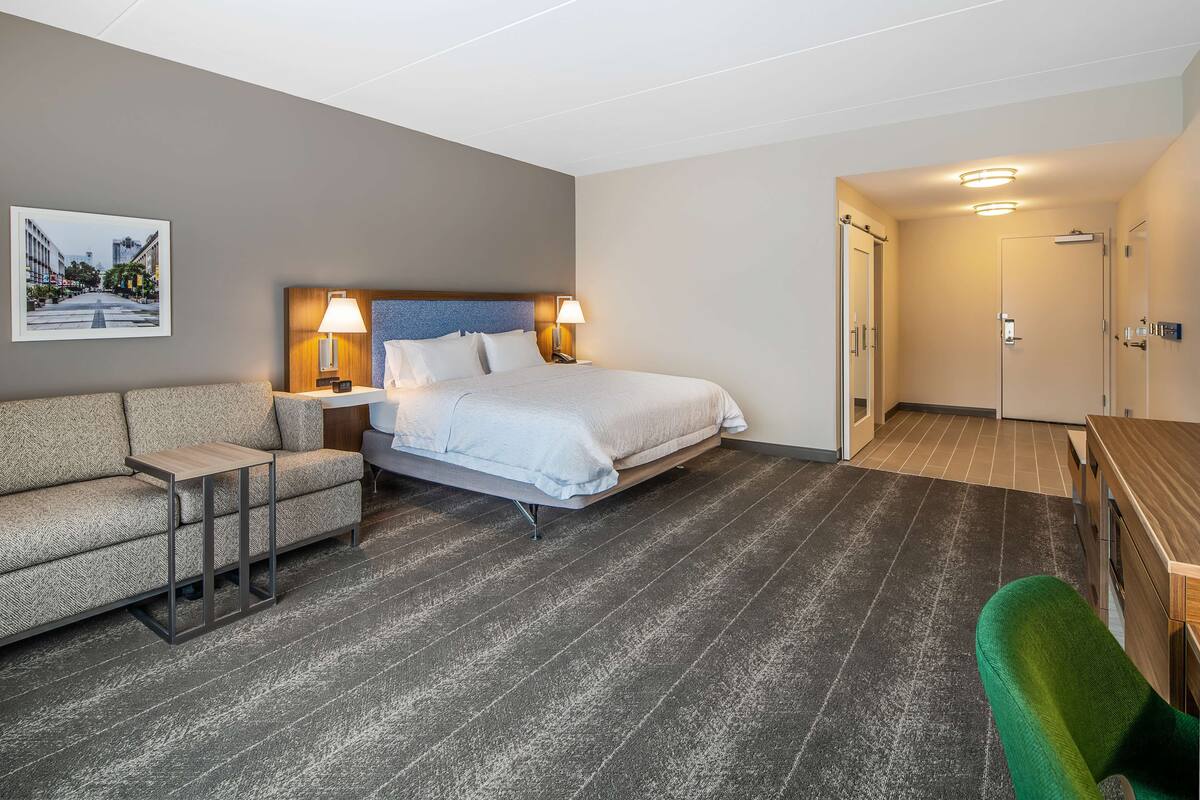 A hotel room with a bed, a sofa, a small table, and a green chair. There is a door leading to a bathroom and a hallway.