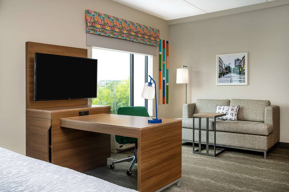 A hotel room with a wooden desk, a flat-screen TV, a green chair, a gray sofa, and a framed picture on the wall.
