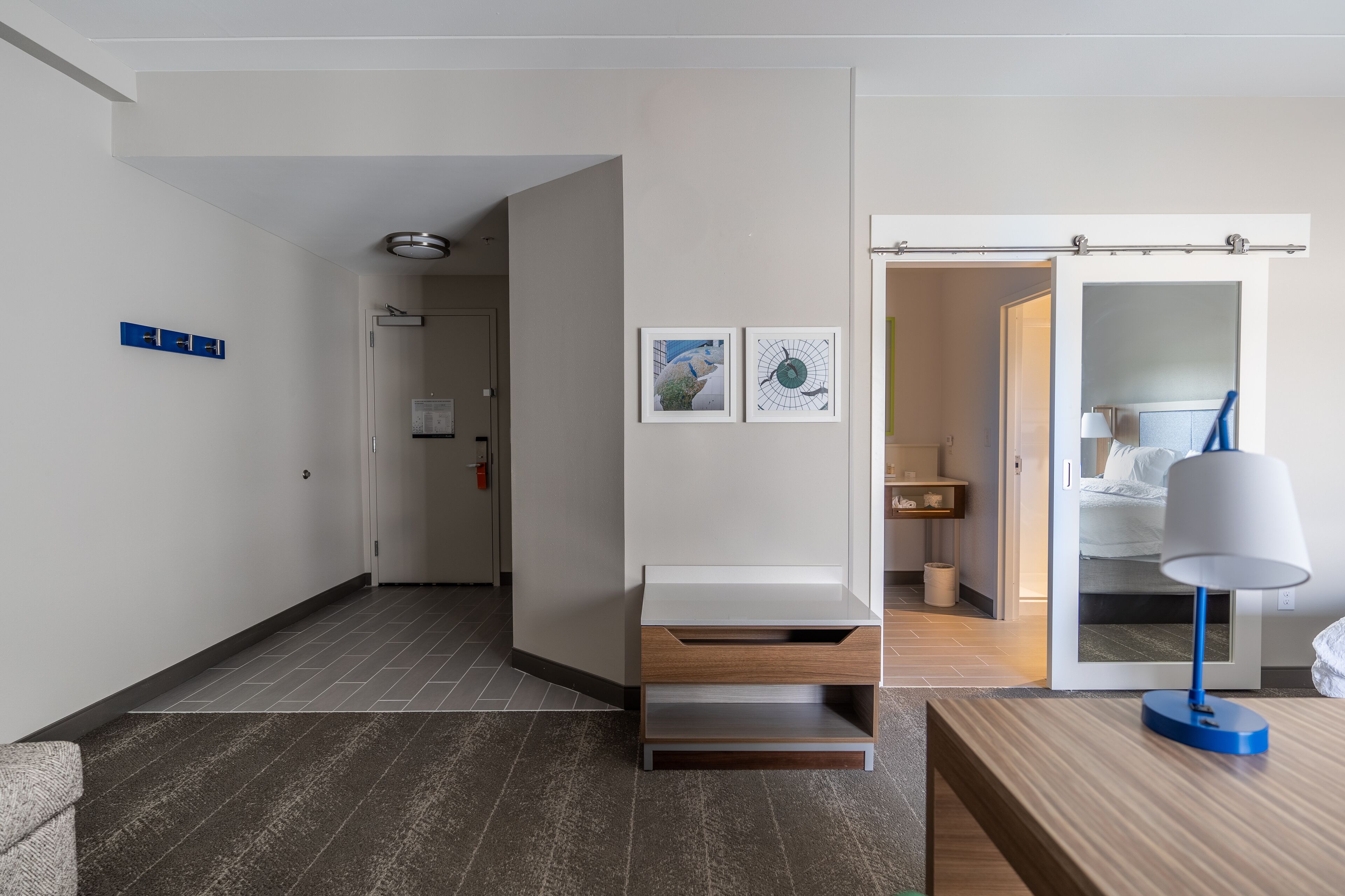 A modern hotel room with a wooden floor, a desk with a lamp, and a bed with a nightstand.