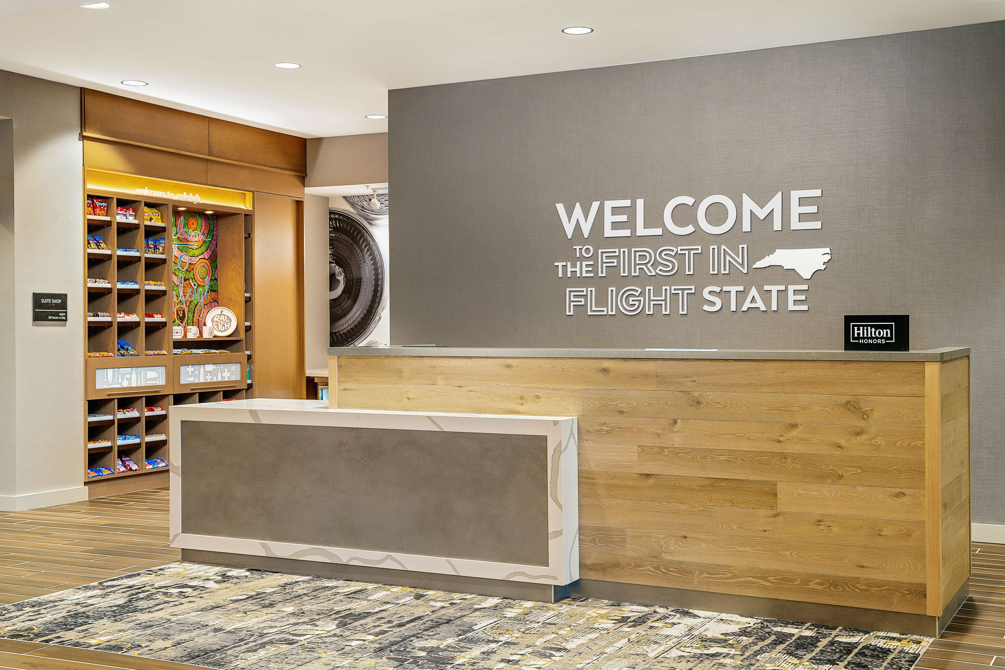 A reception desk with a "WELCOME TO THE FIRST IN FLIGHT STATE" sign, a "Hilton Honors" badge, and a colorful toy display in the background.
