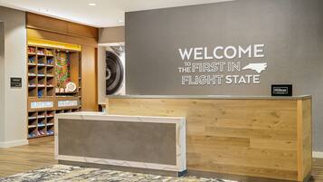 A reception desk with a "WELCOME TO THE FIRST IN FLIGHT STATE" sign, a "Hilton Honors" badge, and a colorful toy display in the background.
