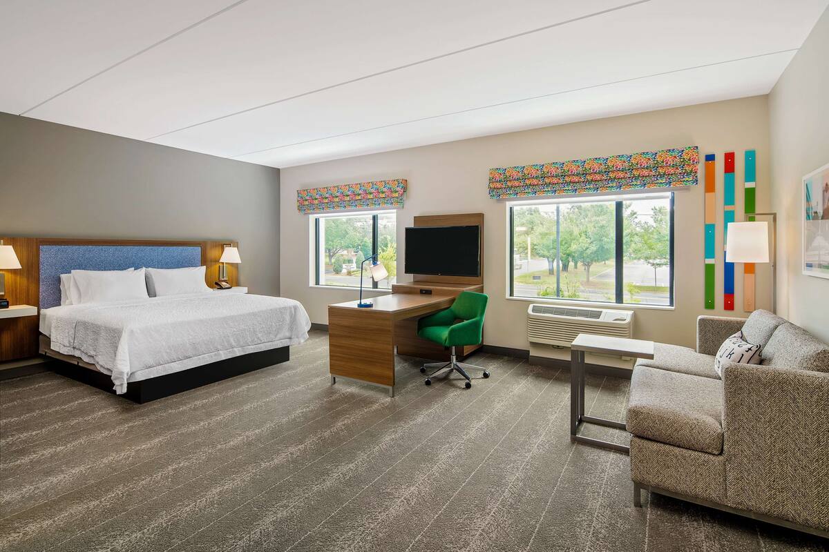 A hotel room with a bed, desk, chair, TV, and large windows.