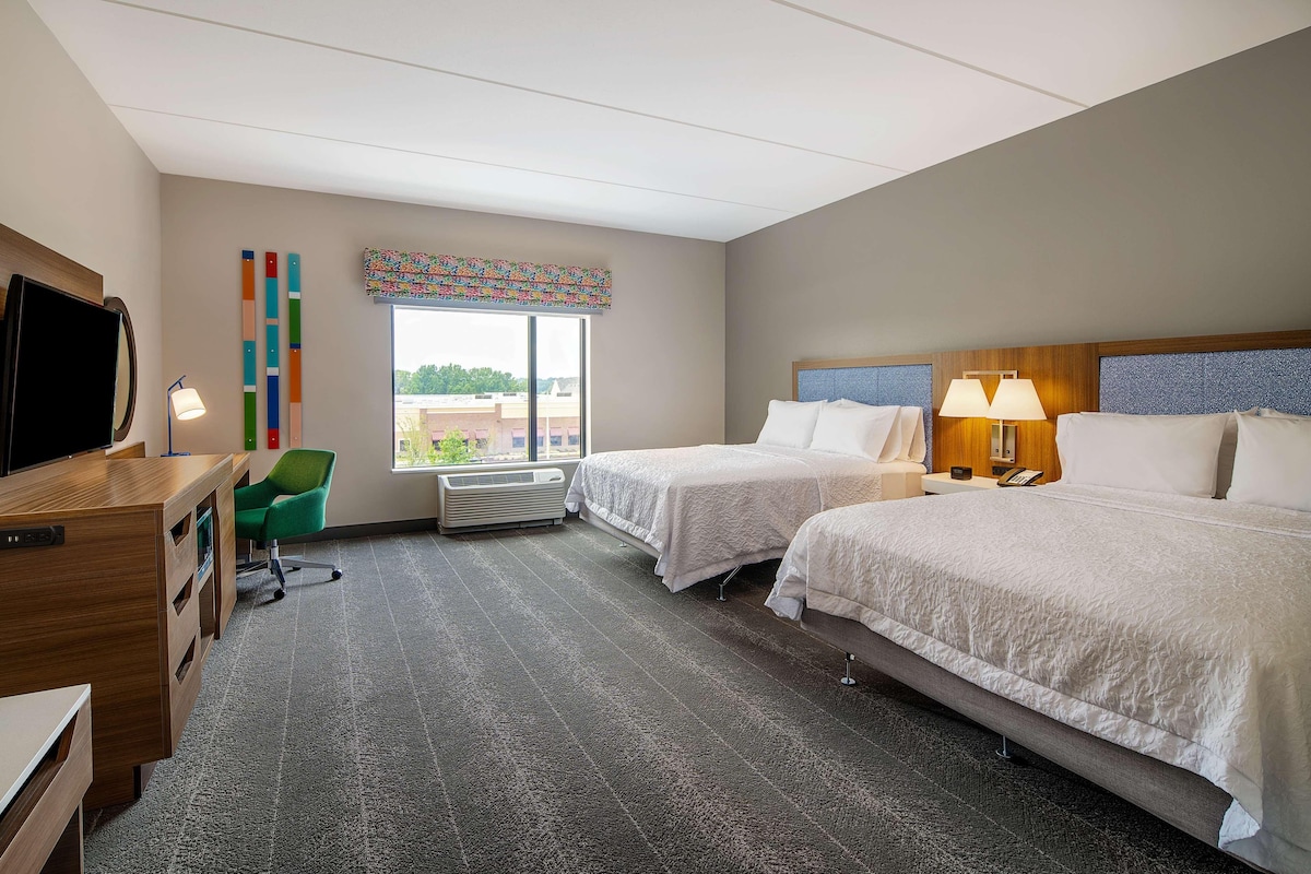 A hotel room with two beds, a desk, a chair, a television, and a window with a view.