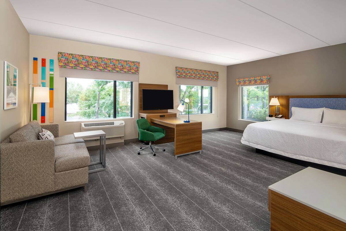 A hotel room with a bed, a desk, a green chair, a TV, and large windows.