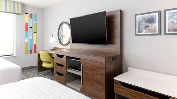 A modern hotel room with a wooden desk, a flat-screen TV, a mirror, and a bed.
