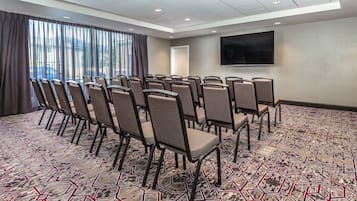 Une salle de conférence avec des rangées de chaises, une grande fenêtre avec des rideaux, un téléviseur à écran plat fixé au mur et une moquette à motifs.
