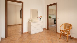 A room with a white dresser, a mirror, a wicker chair, and a kitchen visible through an open doorway. - Experience Italian flair up close - Monolocale 900 (Racale)