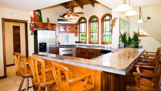A kitchen with wooden bar stools, a large island, and a view of the outdoors through arched windows. - Villa Verde (Sardinal)