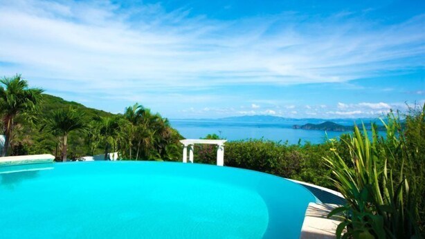 Une piscine d’un bleu éclatant, agrémentée d’une pergola blanche et d’une végétation luxuriante, offrant une vue panoramique sur la mer et les îles lointaines.
