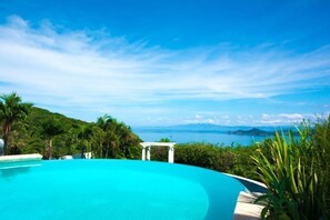 Une piscine d’un bleu éclatant, agrémentée d’une pergola blanche et d’une végétation luxuriante, offrant une vue panoramique sur la mer et les îles lointaines.