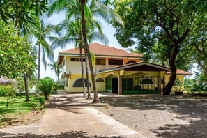A two-story house with a covered porch, surrounded by lush greenery and palm trees.