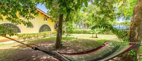 A hammock under a tree in a garden with a yellow building in the background.