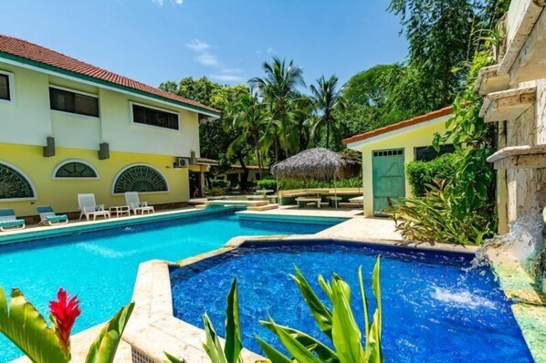 A swimming pool with lounge chairs and a thatched-roof structure, surrounded by lush greenery and a building with arched windows.